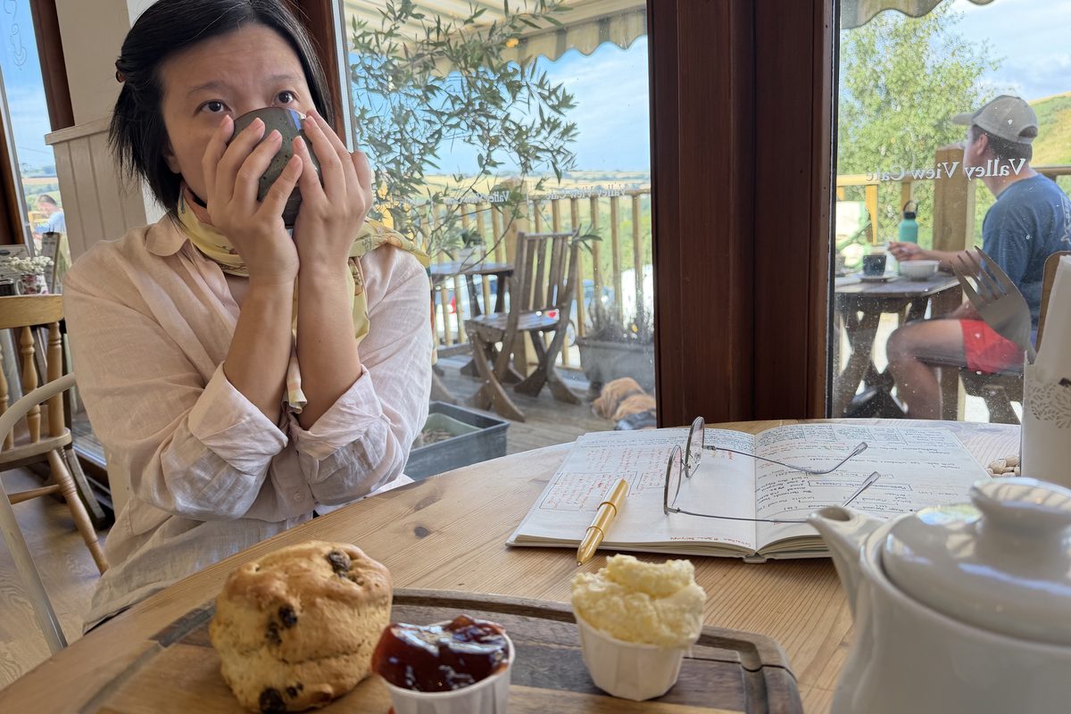 Jern drinking tea beside scones, strawberry jam, clotted cream and her open research notebook at a Devon cafe.
